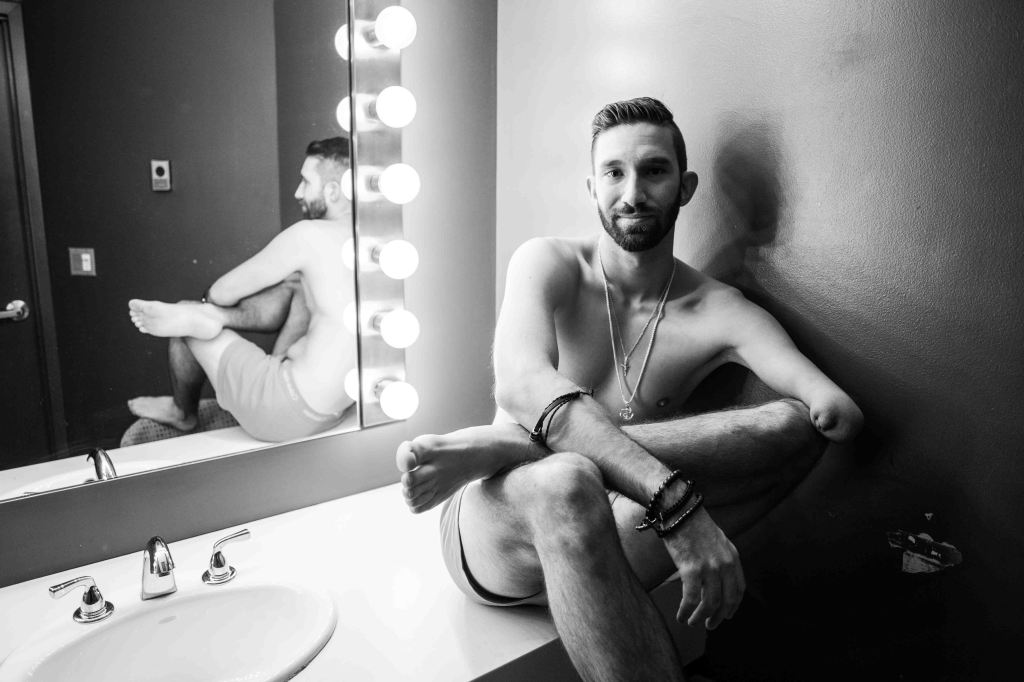 Matthew is sitting on a sink in a studio dressing room. His residual limb is prominently featured, and Matthew is very relaxed.