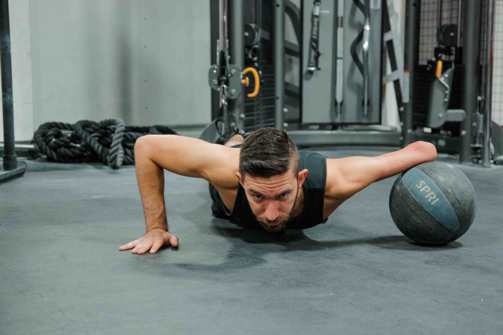 A color photo of Matthew performing a push up. His left residual limb is resting on a medicine ball to allow him to successfully perform the push up with proper form.