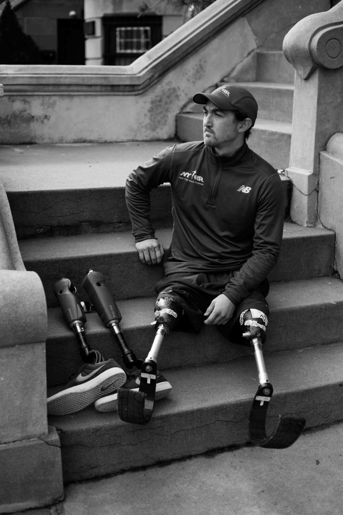 A portrait shot of Rudy on stairs, with his running prostheses on and his walking prostheses beside him. He is composed, and the shot captures Rudy looking at something to the side.