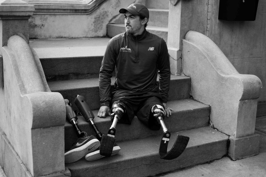 A landscape orientation black and white photo of Rudy sitting on some stairs with his running legs on and his walking prostheses next to him. The shot emphasizes the space around him.