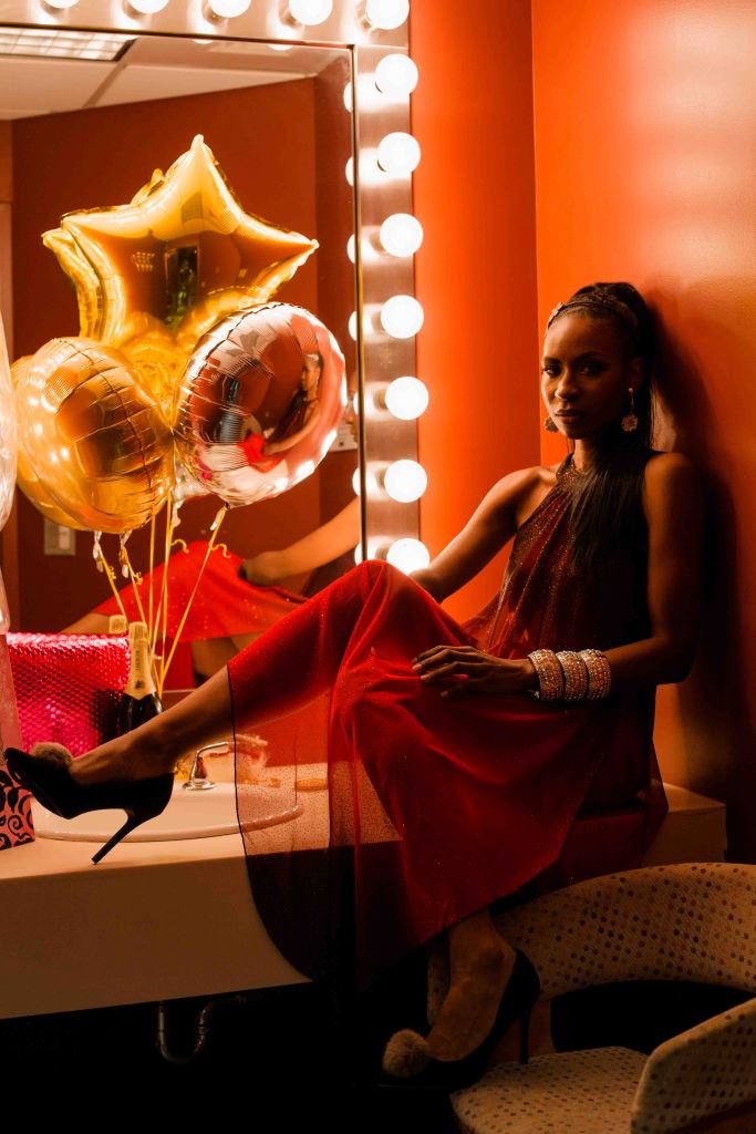 An image of Jaleesa in a studio room, in front of a mirror. There are balloons next to her, including one shaped like a star; her residual limb is obscured by her leg but reflected in the mirror behind her